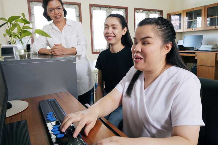 A woman sitting at a desktop computer and navigating using the touch keys of a braille display while two other women smile and look on