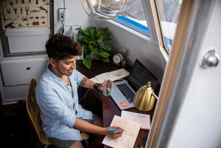 Young worker takes notes at a desk next to their laptop, smiling and holding a glass of water