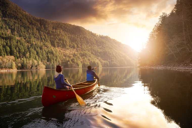 couple in a canoe, paddling up a mountain and tree lined river into the sunset
