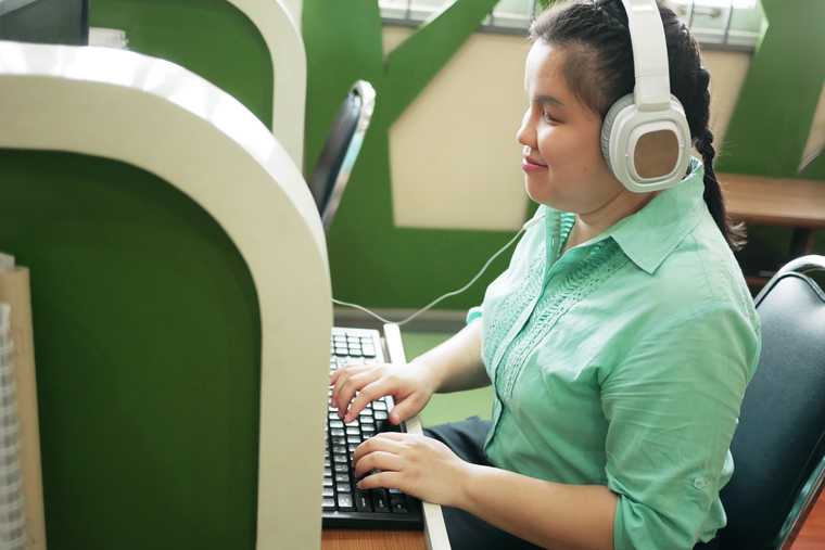 young woman in a cubicle wearing headphones smiles with her hands on a keyboard