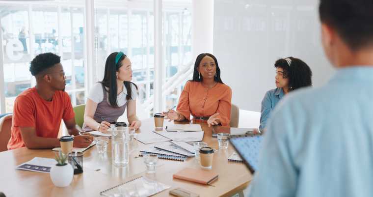 five people around a table with papers spread out, focusing on one woman speaking while gesturing