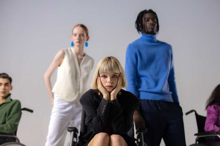 A person with a blond bob haircut wearing a black blazer poses in a wheelchair in front of two people people standing behind them.