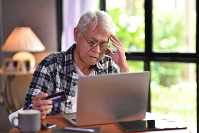Grey-haired man rubs his temple while holding a credit card and staring down at his laptop