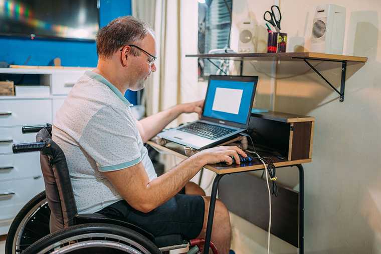 Man sitting in a wheelchair uses a mouse and laptop at a desk setup in a home office