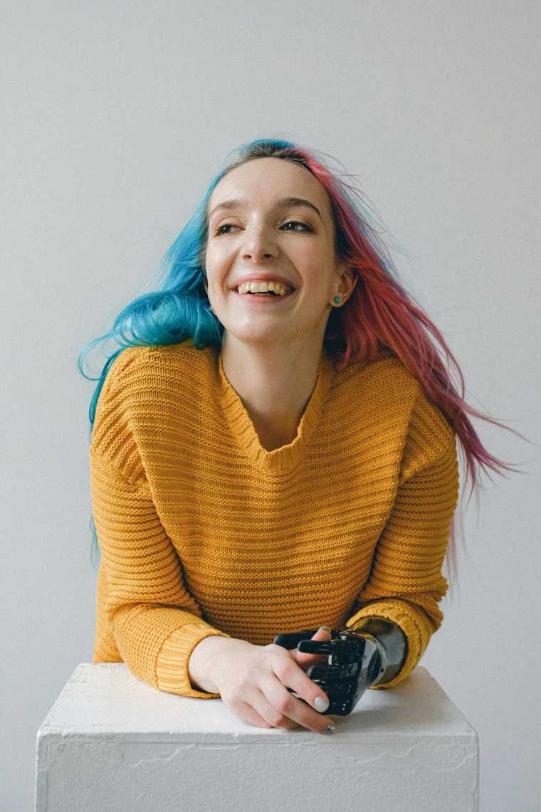 A woman with pink and blue hair, a yellow sweater, and a prosthetic arm poses and smiles while leaning on a white block.