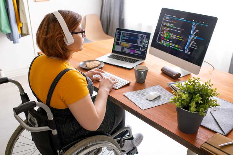 woman seated in wheelchair with headphones at a desk and two monitors both showing code onscreen