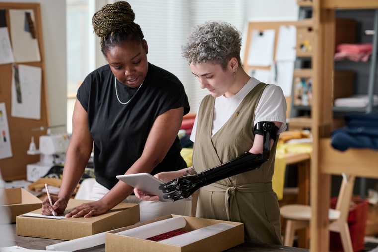 woman with prosthetic arm holds a tablet while speaking with a woman writing on a package