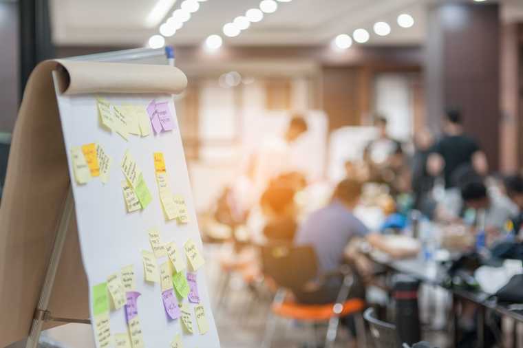 A flipboard covered in sticky notes citing various features, with a boardroom filled with people off in the background