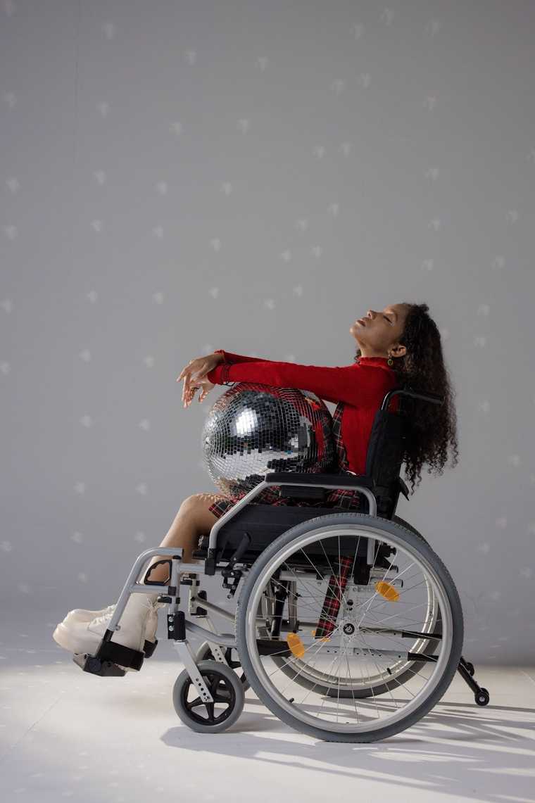 A person in a wheelchair with long, curly hair poses holding a disco wearing a red turtleneck, a plaid overall skirt, and white sneakers.