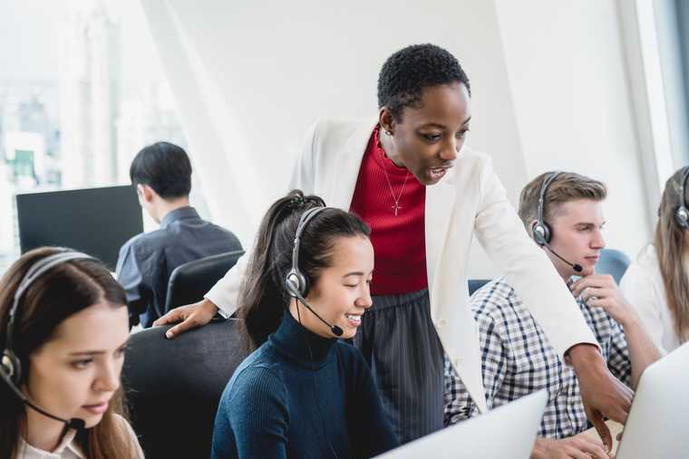 customer support manager stands by an agent's chair, pointing to something on her screen while other agents are on headsets around them